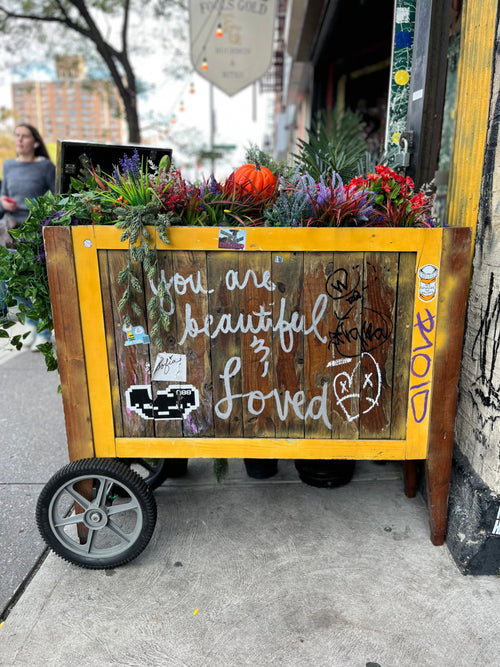 A cart leaning against a wall filled with flowers and a small orange pumpkin with the words "you are beautiful and loved" written on the side. 