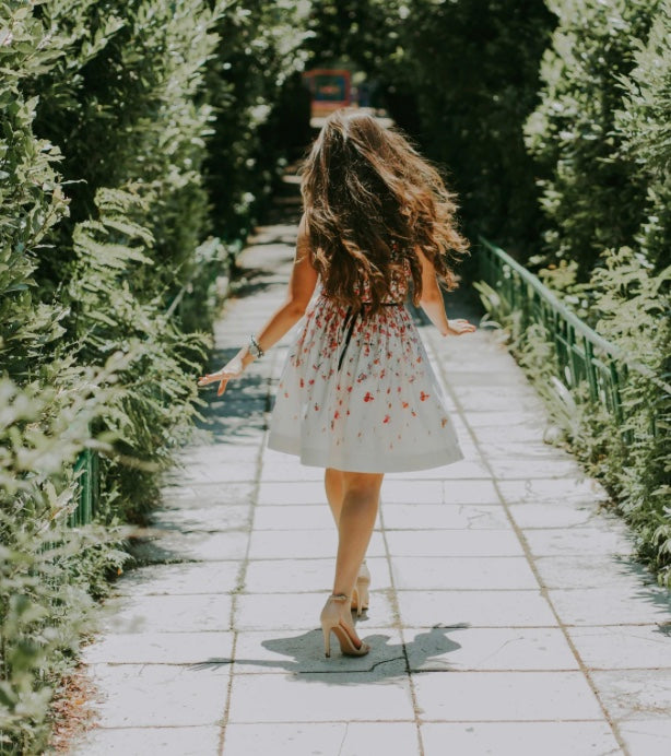 Woman in a floral dress and long hair walking along a path flanked by greenery with her arms swinging at her sides.